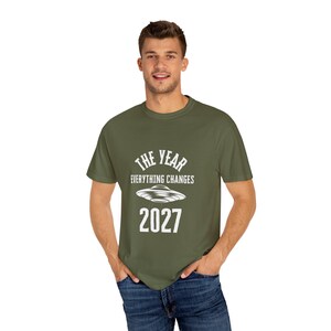 Puede incluir: Camiseta verde oliva con el texto blanco "THE YEAR EVERYTHING CHANGES 2027" y un diseño de OVNI. La camiseta está hecha de un material suave y es adecuada para el uso diario.
