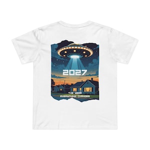 May include: White t-shirt featuring a graphic of a UFO beaming light onto a suburban neighborhood. The design includes the year "2027" and the text "The Year Everything Changes". The artwork is set against a night sky with stars.
