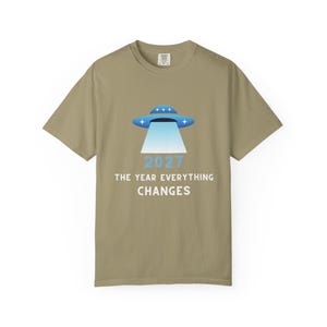 Könnte beinhalten: Khaki-farbenes T-Shirt mit einem blau-weißen UFO-Design und dem Text "2027 THE YEAR EVERYTHING CHANGES". Das Grafik-T-Shirt hat einen Rundhalsausschnitt und kurze Ärmel. Das Shirt ist neutral gefärbt.