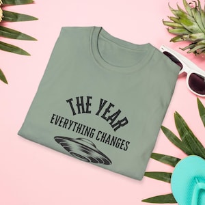 May include: Sage green t-shirt with the text "THE YEAR EVERYTHING CHANGES" and a graphic of a UFO. The shirt is surrounded by tropical leaves, sunglasses, and a flip-flop on a pink background.