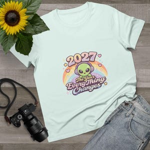 May include: A light blue t-shirt featuring a cartoon alien wearing headphones, set against a rainbow. The shirt has the text "2027 The Year Everything Changes" in a playful font. A sunflower and camera are in the background.