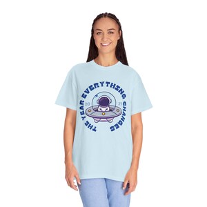 May include: Light blue t-shirt with a circular graphic featuring an astronaut in a UFO. The text around the graphic reads "THE YEAR EVERYTHING CHANGES 2027". The shirt is a comfortable, casual style.