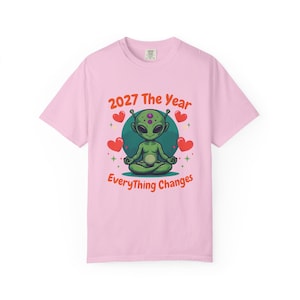 May include: A light pink t-shirt featuring a cartoon alien meditating. The alien is green with purple spots and is surrounded by red hearts and the text "2027 The Year Everything Changes" in red and orange.
