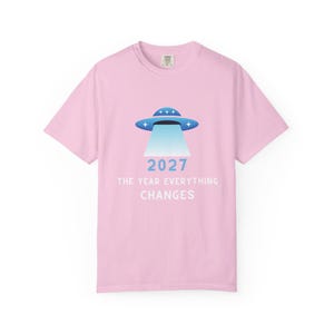 Könnte beinhalten: Hellrosa T-Shirt mit einem blauen und weißen UFO-Design und dem Text "2027 THE YEAR EVERYTHING CHANGES". Das T-Shirt hat einen Rundhalsausschnitt und kurze Ärmel. Das Design ist mittig platziert.