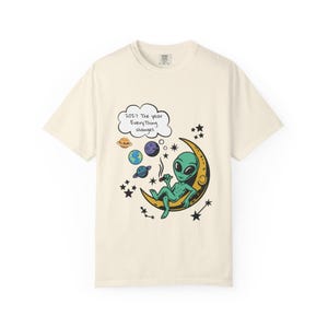 May include: Cream-colored t-shirt featuring a cartoon alien smoking a cigarette while seated on a crescent moon. The design includes planets, stars, and the text "2027 The year Everything changes" in a thought bubble.