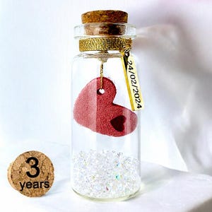 May include: A clear glass bottle with a cork stopper containing a red heart-shaped ornament and small clear crystals. A gold-coloured metal tag hangs from the neck with the text 24/02/2024. A separate cork disc nearby is printed with 3 years.