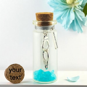 May include: A clear glass bottle with a cork stopper, filled with blue crystals and a silver handcuff charm that reads "FREEDOM." A small tag with the date "30/03/2025" hangs from the bottle. A cork with the text "your text" is on the left.