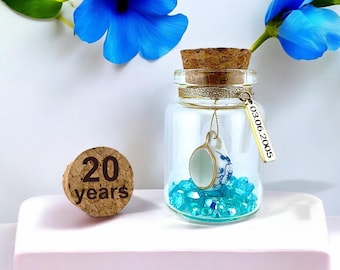 Puede incluir: Un frasco de vidrio transparente con tapón de corcho, que contiene una taza de té de cerámica en miniatura blanca y azul sobre cristales turquesa. Una etiqueta de metal dorado cuelga del cuello, grabada con 03.06.2005. Un corcho aparte dice 20 years.