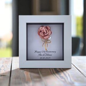 May include: A framed anniversary keepsake. It features a pink fabric rose with a gold bow and pearl accent. The white frame surrounds a black mat with the text "Happy 12th Anniversary Alex & Milana 10.10.2025".