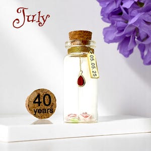 May include: A glass bottle with a cork stopper and a gold ribbon around the neck. Inside the bottle are two small pink flowers and a red teardrop charm hanging from a gold chain. The bottle has a gold tag with the date "05.05.25" written on it. A cork circle with the text "40 years" is in the foreground. The bottle is on a white surface.