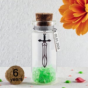 Könnte beinhalten: Eine klare Glasflasche mit Korken, silbernen Akzenten und einem Schwertanhänger. Die Flasche enthält grüne Kristalle und ein Etikett mit dem Datum 05.01.2026. Ein Holzkorken mit der Aufschrift "6 Jahre" ist ebenfalls vorhanden.
