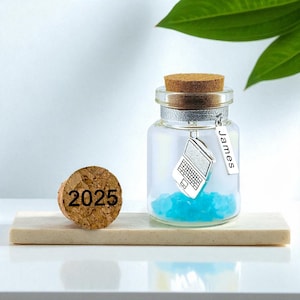 Könnte beinhalten: Eine klare Glasflasche mit Korken, gefüllt mit blauen Dekosteinen und einem silbernen Laptop-Anhänger. Ein Anhänger mit dem Namen "James" hängt an der Flasche. Ein separater Korken mit der Aufschrift "2025" liegt daneben.
