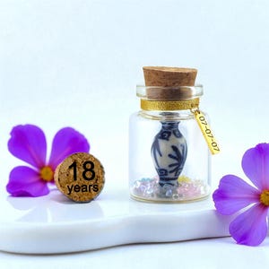 May include: A clear glass jar with a cork stopper, containing a miniature blue and white patterned vase and small colourful beads. A gold ribbon with a rectangular tag reading 07-07-07 is tied around the neck. A separate cork reads 18 years.