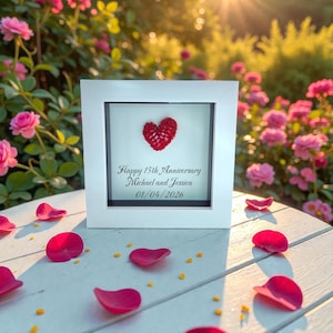 Könnte beinhalten: Ein weiß gerahmtes Jubiläumsgeschenk mit einem roten Herzdesign. Der Text lautet "Happy 15th Anniversary Michael and Jessica 01/04/2026". Rosa Rosenblätter sind auf einer weißen Oberfläche verstreut.