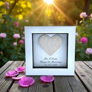 May include: A white framed heart-shaped artwork with a lace design. The inscription reads "Alex & Emilia Happy 13th Anniversary 02/02/2026". Pink flower petals are scattered on a wooden surface in front of the frame.