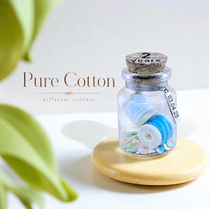 May include: A clear glass jar with a cork stopper labeled 2 years, containing two small spools of light blue cotton thread and scattered pastel heart-shaped confetti. A silver metallic tag hangs from the neck, printed with 03.04.25.