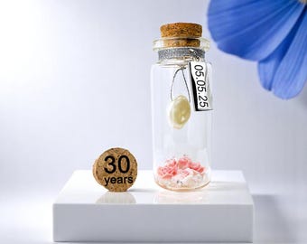 May include: A small glass bottle with a cork stopper and a silver tag that says "05.05.25". Inside the bottle are pink and white flowers and a white pearl. A cork stopper with the text "30 years" is in the foreground.