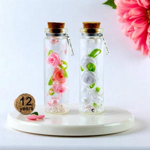Peut inclure: Deux bouteilles en verre avec bouchons en liège, remplies de roses en tissu roses et blanches, de perles et de cristaux. Chaque bouteille a une étiquette argentée avec une date. Un bouchon avec "12 ans" est à côté.