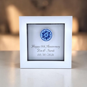 May include: A square white shadow box frame containing a circular blue and white floral ceramic piece. The glass features black printed text that reads: Happy 18th Anniversary, Tom & Sarah, 08/10/2026. The frame has a clean, simple border design.