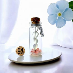 May include: A clear glass bottle with a cork stopper, containing heart-shaped beads and pink decorative elements. A small metal tag displays the date "05.05.25". A cork with "24 years" is next to the bottle. A blue flower is in the background.