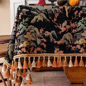 Może przedstawiać: Czarny obrus z szczegółowym, inspirowanym Azją wzorem krajobrazu z górami, drzewami i jeleniami. Obrus jest obszyty złotą lamówką i ozdobnymi chwostami w odcieniach pomarańczu i bieli.