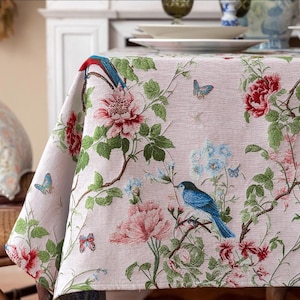 May include: A pink tablecloth with a floral pattern featuring blue birds, butterflies, and green leaves. The tablecloth has a subtle texture and a delicate design.