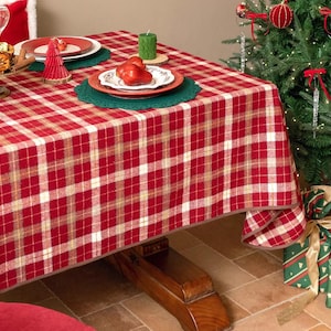 Op de afbeelding: Een rood-wit geruite tafelkleed bedekt een houten tafel die is gedekt voor een feestelijke maaltijd. Borden, glazen en een decoratieve kerstboom zijn zichtbaar. Op de achtergrond staat een kerstboom met versieringen en ingepakte cadeaus.