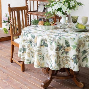 May include: A round tablecloth with a botanical leaf pattern in shades of green, brown, and yellow covers a wooden table. The table is set with plates, glasses, and a centerpiece of flowers and fruit. A wooden chair is positioned to the side.