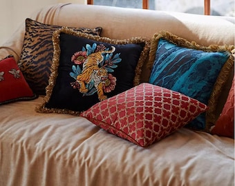 May include: A beige sofa with five throw pillows. The pillows are decorated with various patterns, including a tiger, a geometric design, and a blue and black marbled pattern. The pillows have gold fringe trim.