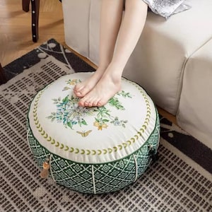 Könnte beinhalten: Runder Hocker mit floralem Druck, mit einer weißen Oberseite mit blauen und grünen Blumen und einem Schmetterling. Die Seiten sind grün mit einem geometrischen Muster. Eine dekorative Quaste hängt an der Seite. Der Hocker steht auf einem gemusterten Teppich.