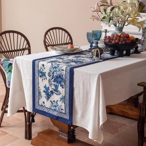 May include: A white tablecloth with a blue floral patterned table runner. The runner features a detailed design with blue flowers and foliage. The table is set with a bowl of grapes, glasses, and a vase of flowers. Two wooden chairs are visible.
