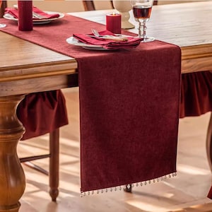 May include: A burgundy velvet table runner with a beaded fringe, set on a wooden table. The table is set with plates, napkins, silverware, and candles, creating an elegant dining scene. The runner adds a touch of luxury.