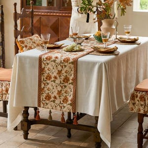 Könnte beinhalten: Ein Esstisch, der für eine Mahlzeit gedeckt ist. Der Tisch ist mit einer cremefarbenen Tischdecke und einem floralen Tischläufer bedeckt. Der Läufer hat einen braunen Rand und Quasten. Stühle mit passenden Kissen umgeben den Tisch. Geschirr, Gläser und eine Vase mit Blumen stehen auf dem Tisch.