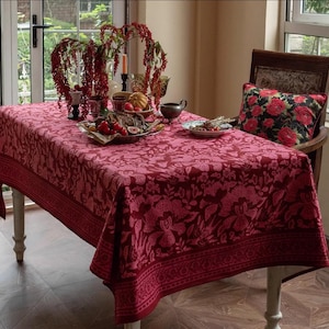 Può includere: Una tovaglia da tavola rossa a fiori con un bordo bianco, posta su un tavolo con una sedia e un cuscino floreale. La tovaglia presenta un motivo floreale tradizionale ed è perfetta per una sala da pranzo o una cucina.
