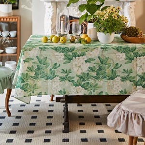May include: A green and white floral tablecloth with a white background and green leaves. The tablecloth is laid on a black and white chequered floor. There is a table with a vase of flowers and a bowl of fruit on top of the tablecloth.