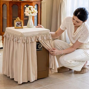 May include: A beige fabric cover for a safe, featuring a pleated skirt with decorative tassels along the top edge. The cover is draped over a rectangular brown safe, with a small gold-toned picture frame and a white vase of flowers on top.