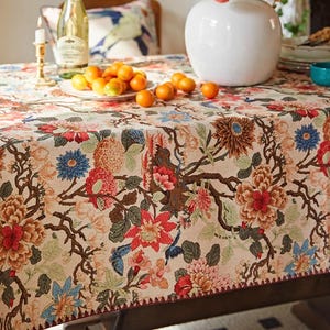 May include: A beige floral tablecloth featuring a detailed pattern of red, blue, and orange flowers and brown branches. The tablecloth is on a wooden table, with a white vase, a plate of oranges, and a bottle of sparkling wine.