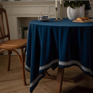 Può includere: Una tovaglia rotonda blu scuro con un bordo di pizzo bianco. La tovaglia è drappeggiata su un tavolo di legno con una sedia sullo sfondo.