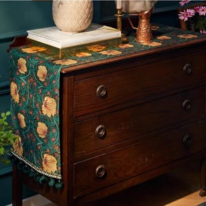 May include: A dark wooden dresser with four drawers, topped with a green floral runner. The runner features a repeating pattern of yellow flowers and red accents. A decorative vase and books sit on top. The runner has a fringe with green tassels.