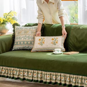 May include: A green sofa with floral patterned cushions and a matching skirt. The cushions feature botanical designs in yellow, green, and white. A person is holding one of the cushions. A teacup and book are on the sofa.