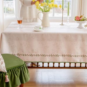 May include: A dining table set for a meal. The table is covered with a light pink floral tablecloth with a green border. A green chair with a ruffled skirt is in the foreground. A vase of yellow flowers, a teacup, and a fruit bowl are on the table.