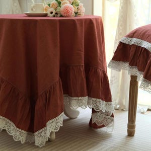 May include: A round, terracotta-coloured tablecloth with a ruffled edge and white lace trim. The tablecloth is draped over a table, with a matching chair cover visible. A cup and flowers are on the table.