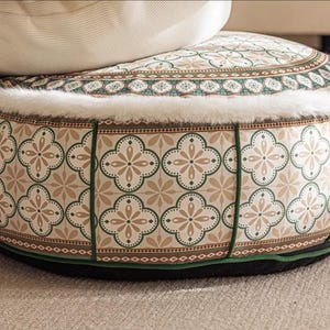 Può includere: Un pouf rotondo con motivo verde e marrone con un bordo in pelliccia sintetica bianca. Il pouf ha una base nera.