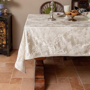 Op de afbeelding: Een wit damast tafelkleed met een delicate kantversiering, gedrapeerd over een houten tafel met een gegraveerd poot. Het tafelkleed heeft een subtiel bloemenpatroon en is gedekt met een zilveren serveerschaal en een glazen kan.