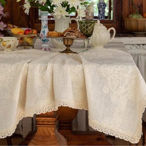 May include: A cream-coloured lace tablecloth featuring detailed floral designs and a scalloped lace border. The tablecloth is draped over a wooden table, creating a vintage and elegant aesthetic. The table is set with a variety of decorative items, including a teacup, flowers, and a cake stand.