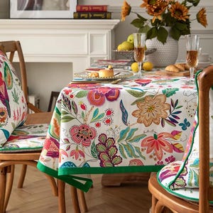 Op de afbeelding: Een eettafelset met een bloemen tafelkleed en bijpassende stoelkussens. Het tafelkleed heeft een witte achtergrond met kleurrijke bloemen en groene rand. De stoelen zijn van hout met kussens met een vergelijkbaar bloemenpatroon.