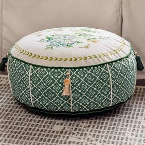 Könnte beinhalten: Ein runder, gepolsterter Ottoman mit weißem Blumendekor und grün gemusterten Seiten. Die Oberseite hat einen botanischen Druck mit blauen und gelben Akzenten. Grüne Quasten und Zierleisten sorgen für Details.