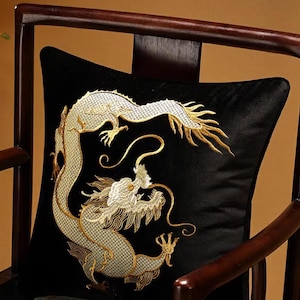 May include: Black square pillow with an embroidered gold dragon design. The dragon is intricately detailed with a white head and gold accents. The pillow is placed on a dark wooden chair, creating a contrast.