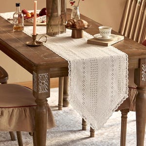 Peut inclure: Un chemin de table en crochet blanc avec un motif de dentelle délicat, posé sur une table en bois avec une chaise brune. Le chemin de table mesure environ 70 cm de large et 180 cm de long.