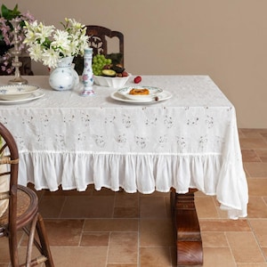 May include: A white floral tablecloth with a frilled edge adorns a wooden table. The table is set with plates, a vase of white flowers, and a decorative candle holder. The scene suggests a refined and elegant dining experience.